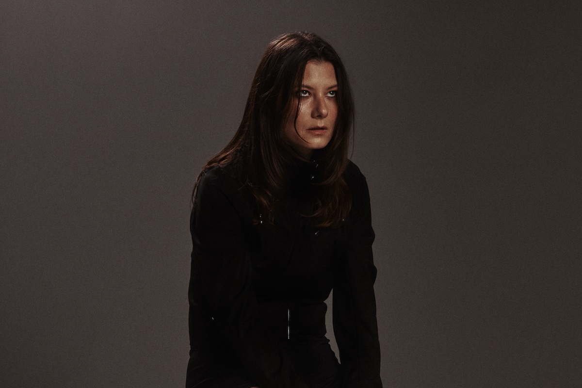 Kate "Babyshakes" Dillon of Full Flower Moon Band in a long-sleeved black outfit, posing against a dark background.